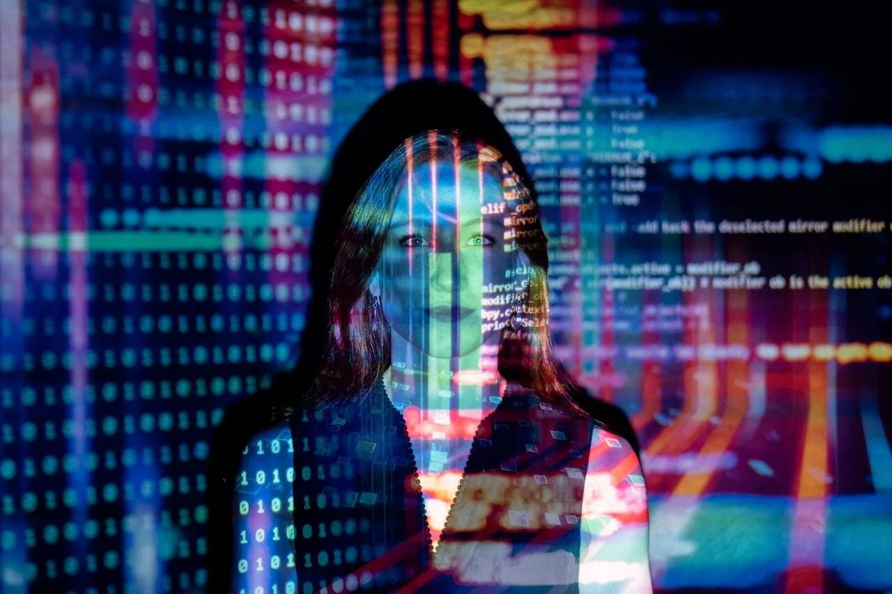 Mujer joven con código binario y líneas de programación proyectadas sobre su rostro y cuerpo, en entorno tecnológico colorido.