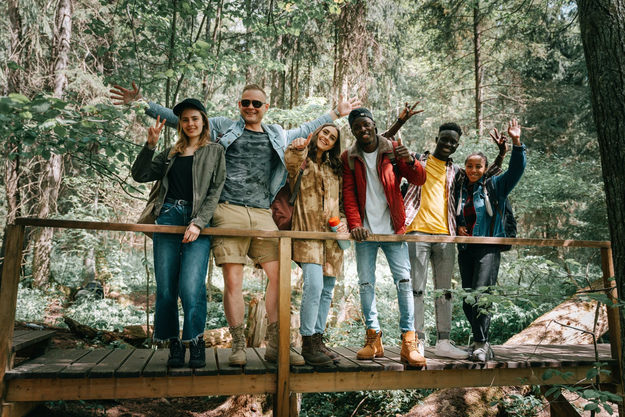 Seis jóvenes diversos posan sonriendo y saludando desde un puente de madera rodeado de árboles durante una excursión.