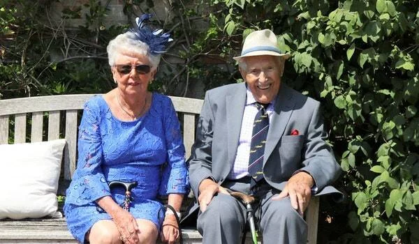 Pareja de ancianos elegantemente vestidos sonríen sentados en un banco de madera en un jardín soleado.