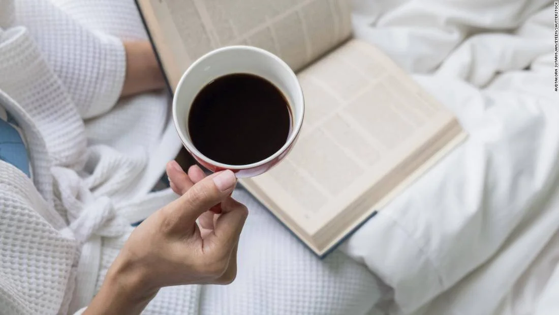 Mano sosteniendo una taza de café negro mientras lee un libro abierto en la cama, con bata blanca.