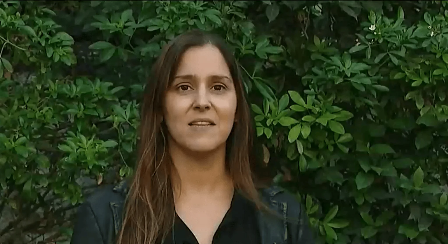 Mujer de cabello castaño largo habla frente a una pared de arbustos verdes.