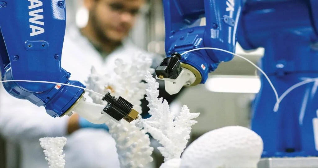 Dos brazos robóticos industriales azules fabrican una estructura tridimensional con forma de coral blanco, supervisados por un técnico al fondo.