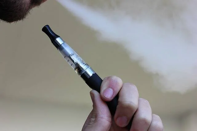 Una persona sostiene un cigarrillo electrónico mientras exhala vapor visible al fondo.