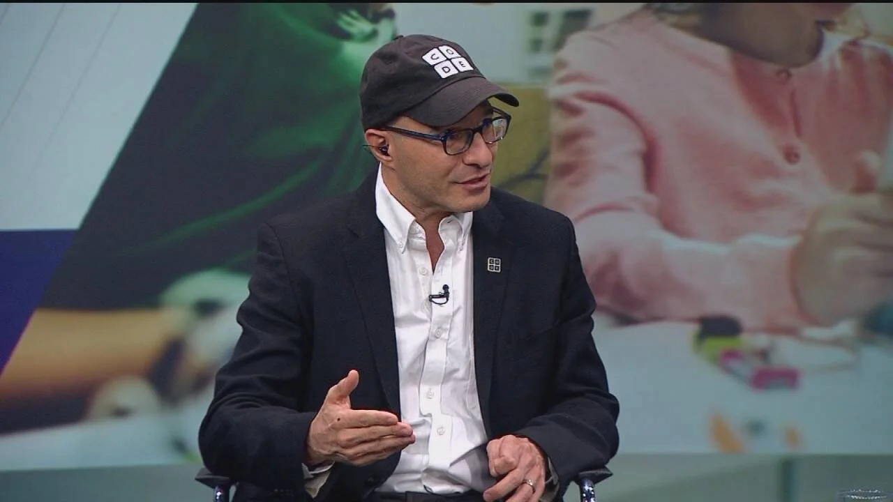 Hombre con gorra oscura, gafas y blazer negro habla en un estudio de televisión con micrófono de solapa.