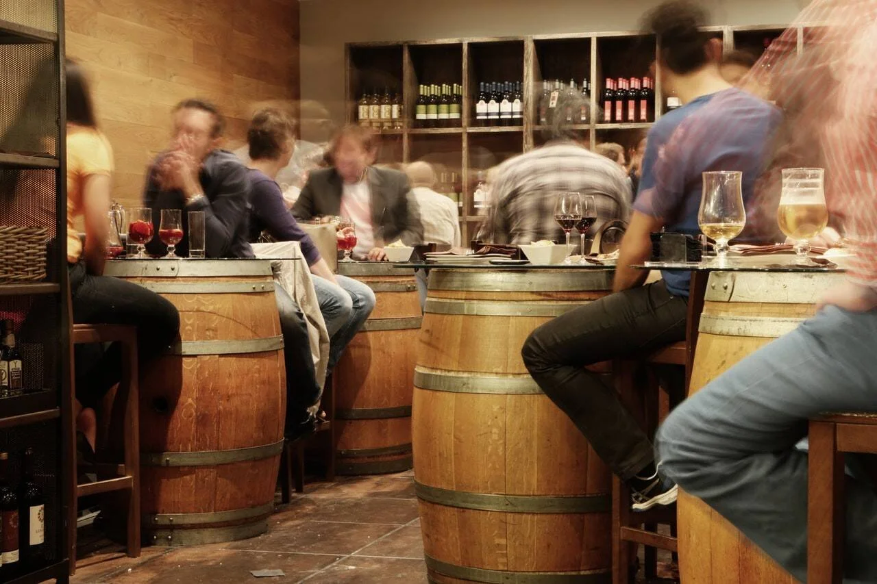 Clientes sentados alrededor de barriles de madera usados como mesas en un bar animado con botellas de vino en estantes al fondo.