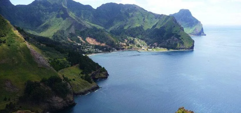 Vista aérea de una bahía tranquila rodeada de montañas verdes con un pequeño poblado costero al fondo.