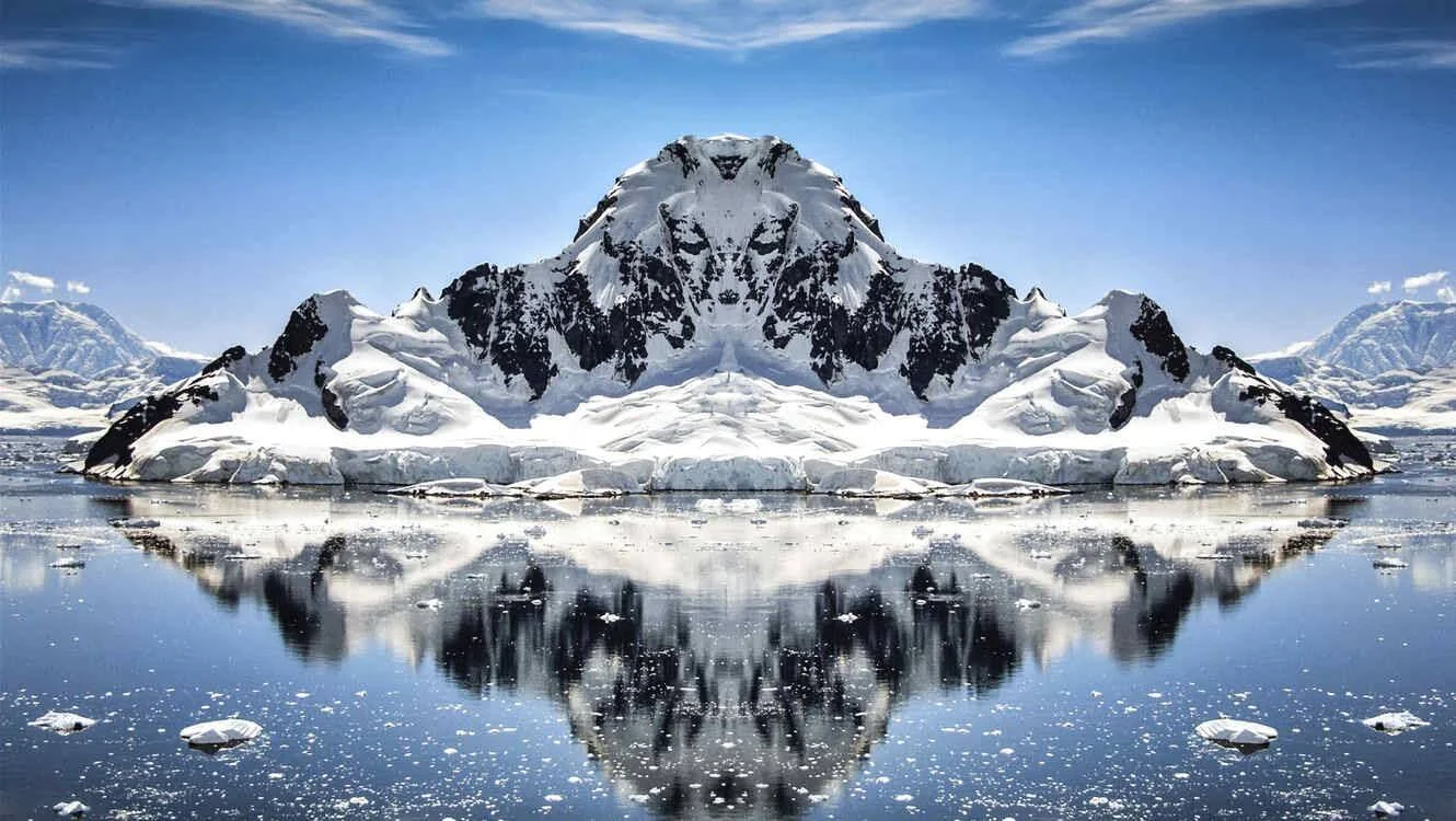 Composición simétrica de una montaña nevada en paisaje polar, con su reflejo perfecto en aguas heladas bajo cielo azul.
