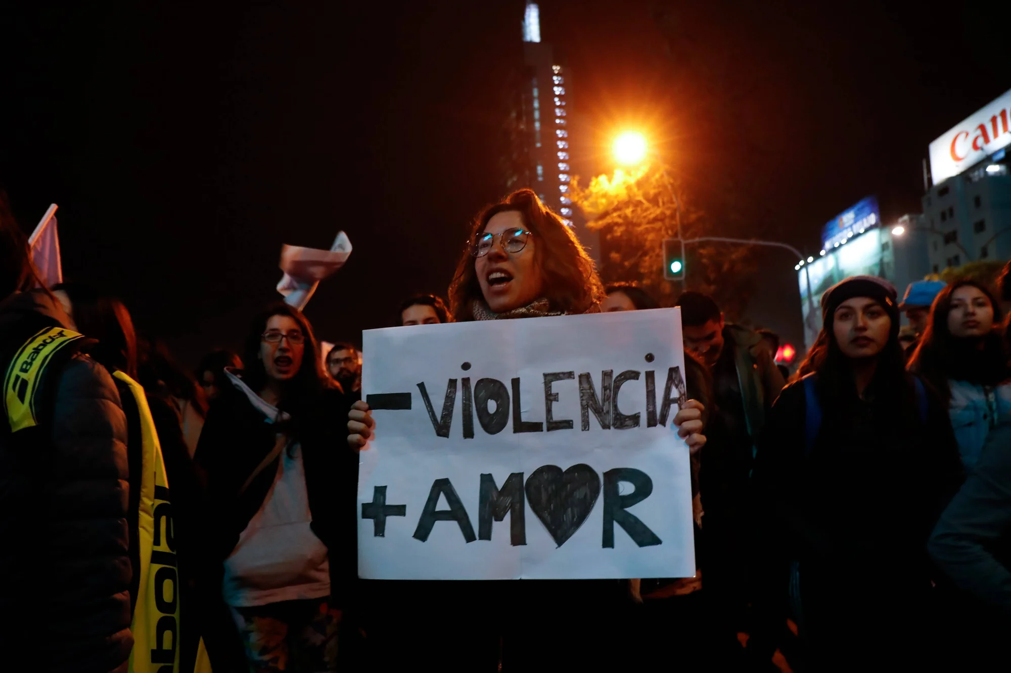 Mujer sostiene un cartel que dice 'Menos violencia, más amor' durante una manifestación nocturna en la calle.