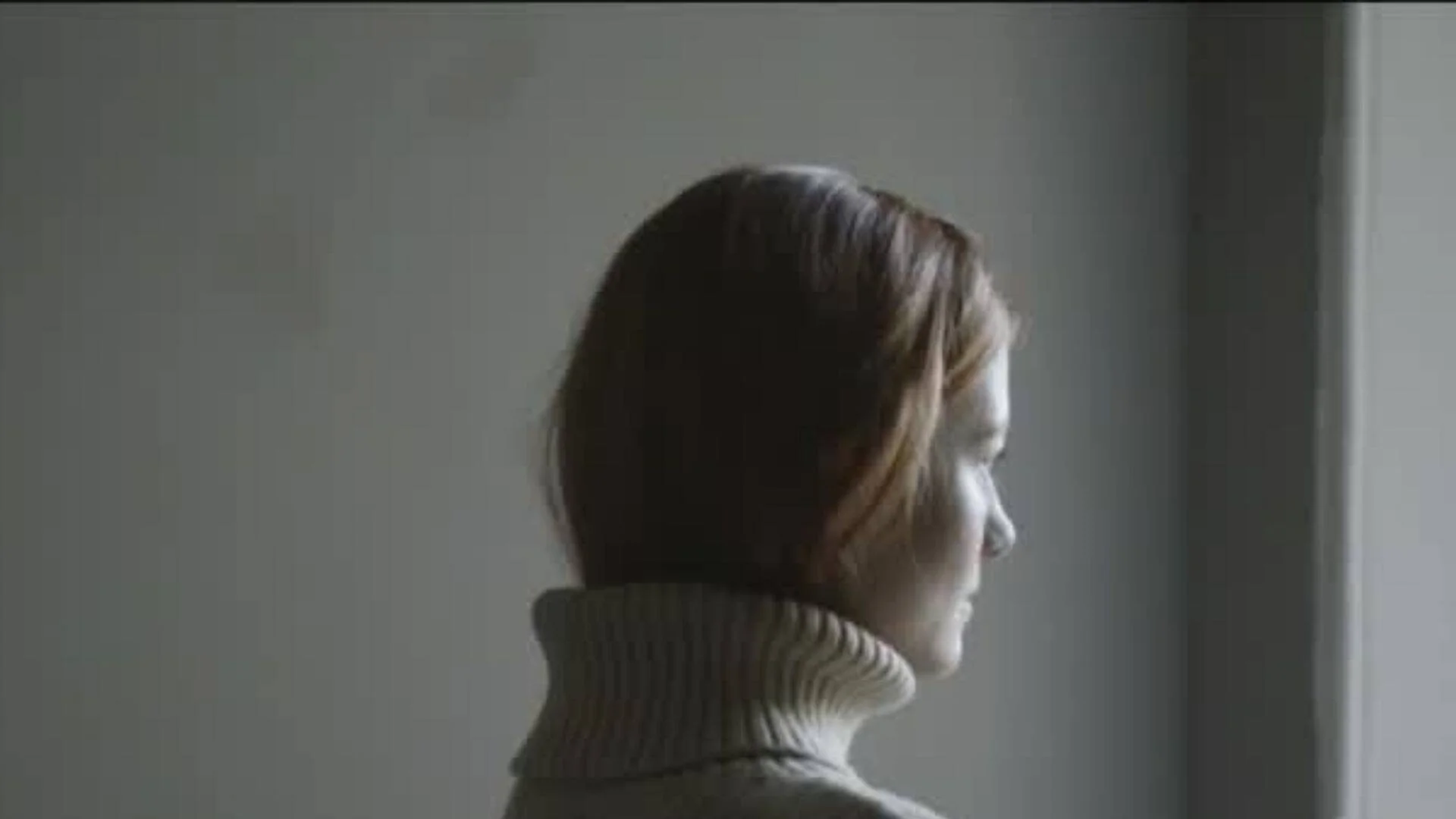 Mujer joven de espaldas, con suéter de cuello alto, mirando de perfil en una habitación tenuemente iluminada.