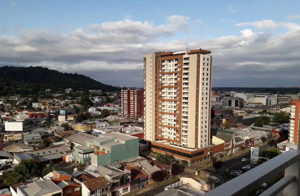 Vista aérea de una ciudad con edificio de gran altura en el centro, rodeado de construcciones de baja altura y un cerro al fondo.