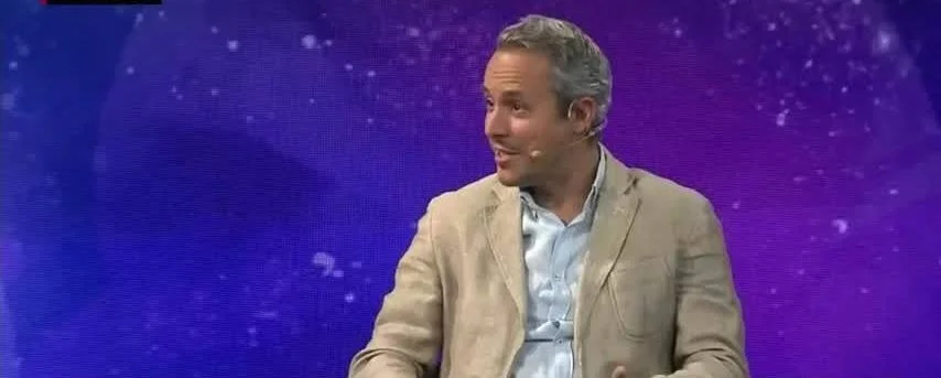 Hombre de cabello canoso con saco beige y micrófono de solapa sonriendo en un estudio de televisión con fondo violeta.