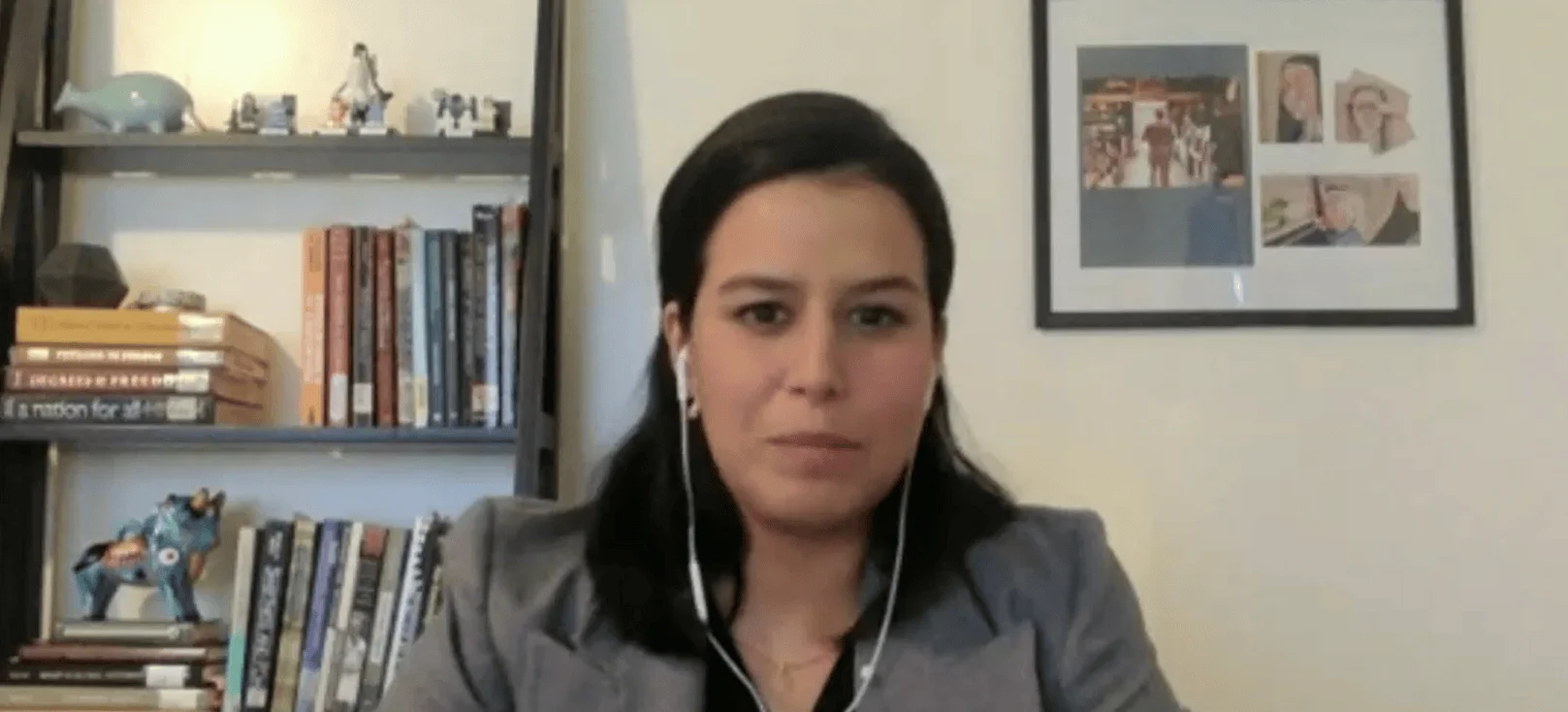 Mujer de cabello oscuro con auriculares blancos participa en una videollamada desde una oficina con estantes de libros al fondo.