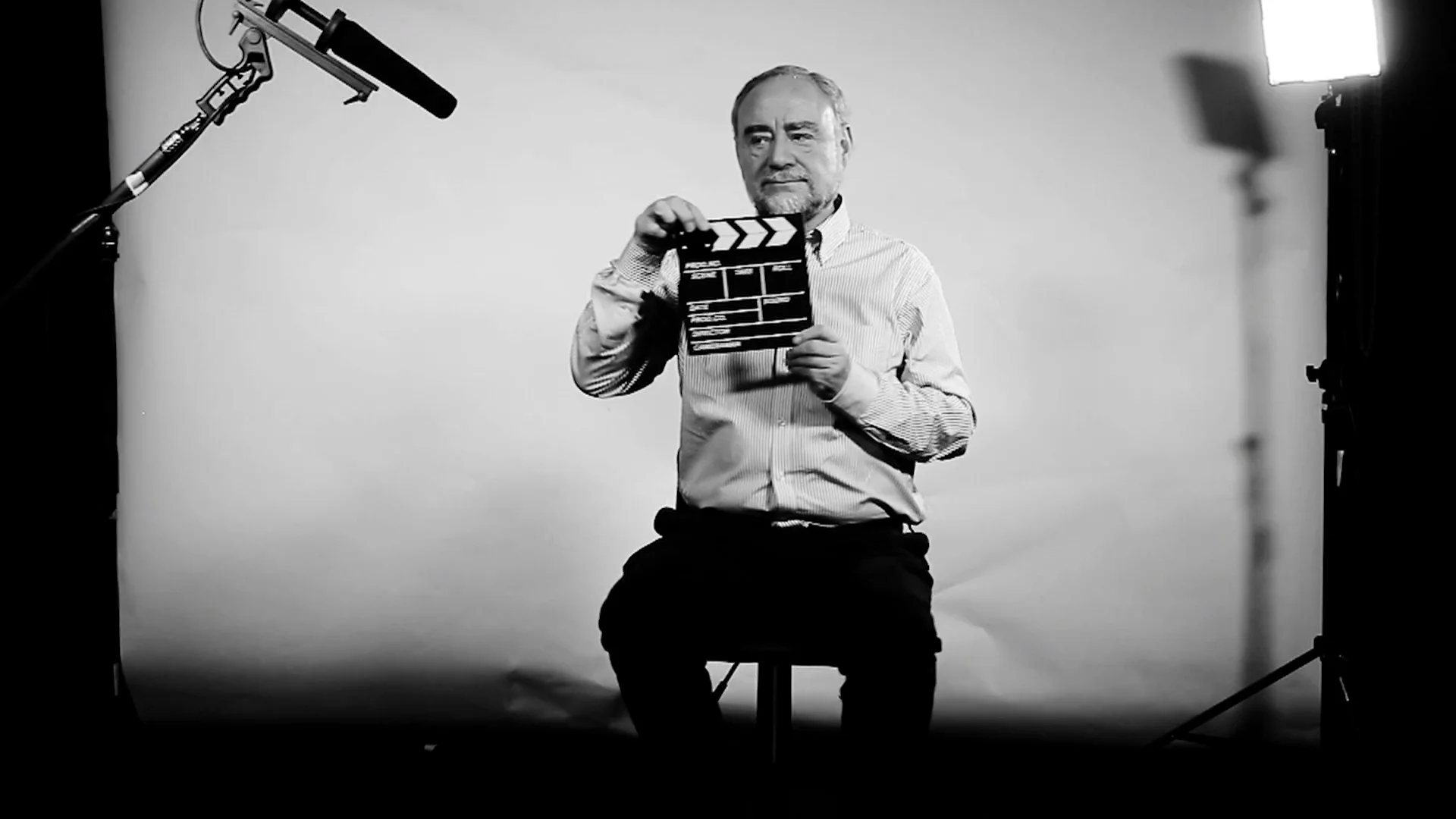 Hombre mayor sentado sostiene una claqueta de cine frente a cámara en un set con micrófono boom y luces, en blanco y negro.