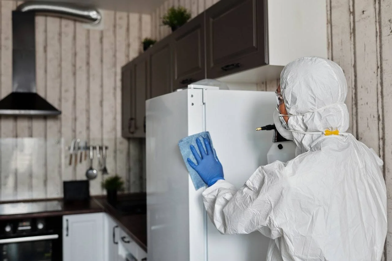 Persona con traje blanco de protección, mascarilla y guantes azules limpia y desinfecta un refrigerador en una cocina.