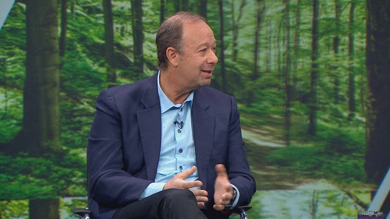 Hombre de mediana edad con saco azul marino y camisa celeste habla gesticulando en un estudio de televisión con fondo de bosque.
