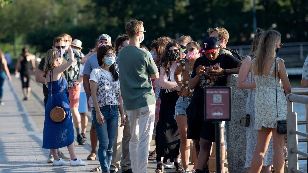 Numerosas personas, varias con mascarilla, hacen fila en un paseo junto al agua durante la pandemia de COVID-19.