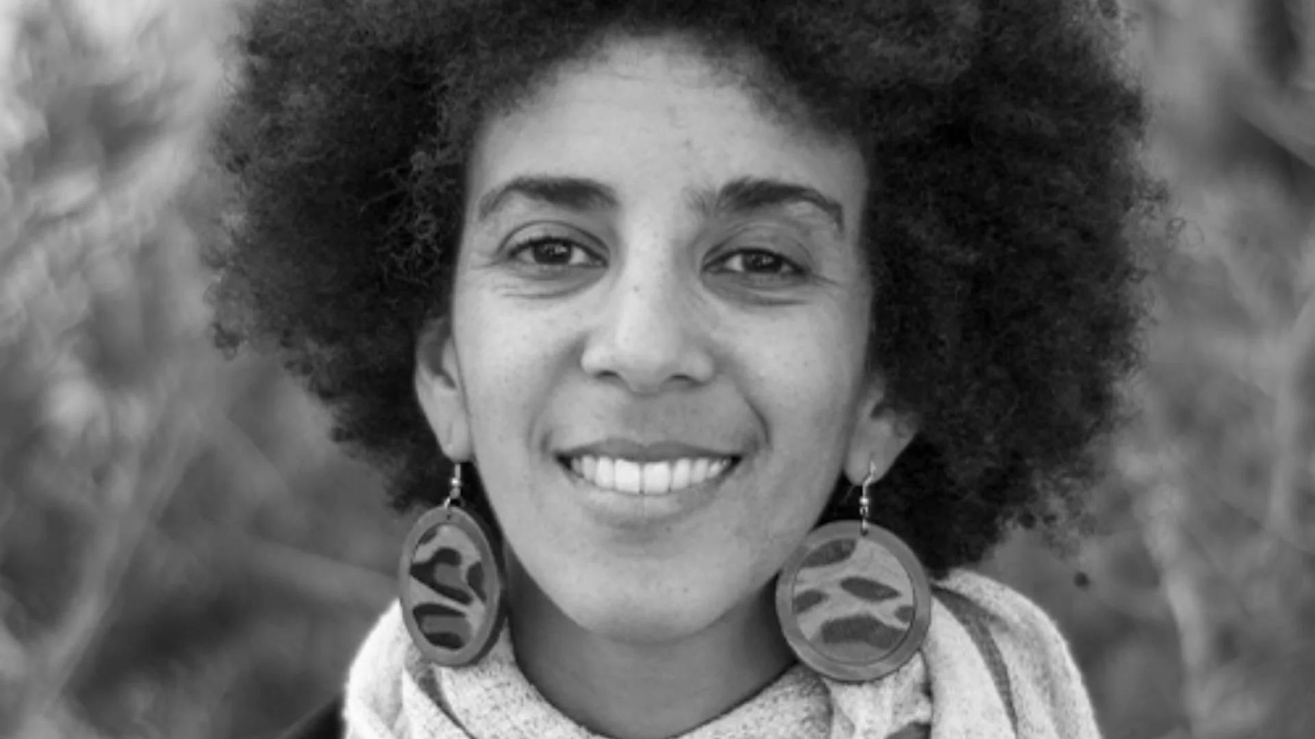 Mujer joven con cabello afro y aretes grandes circulares sonríe a cámara en foto en blanco y negro.