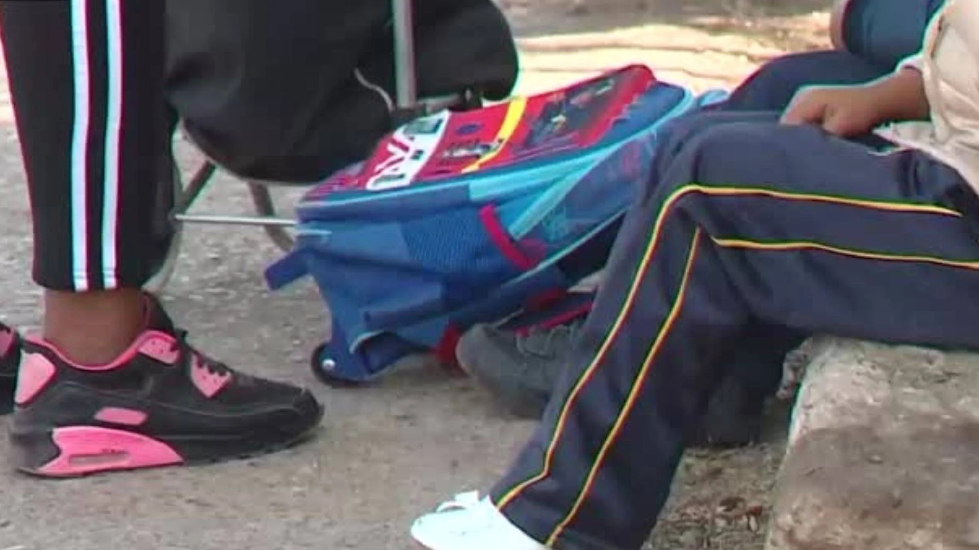 Piernas de menores con mochilas escolares sentados en la calle, con uniformes deportivos y zapatillas de colores.