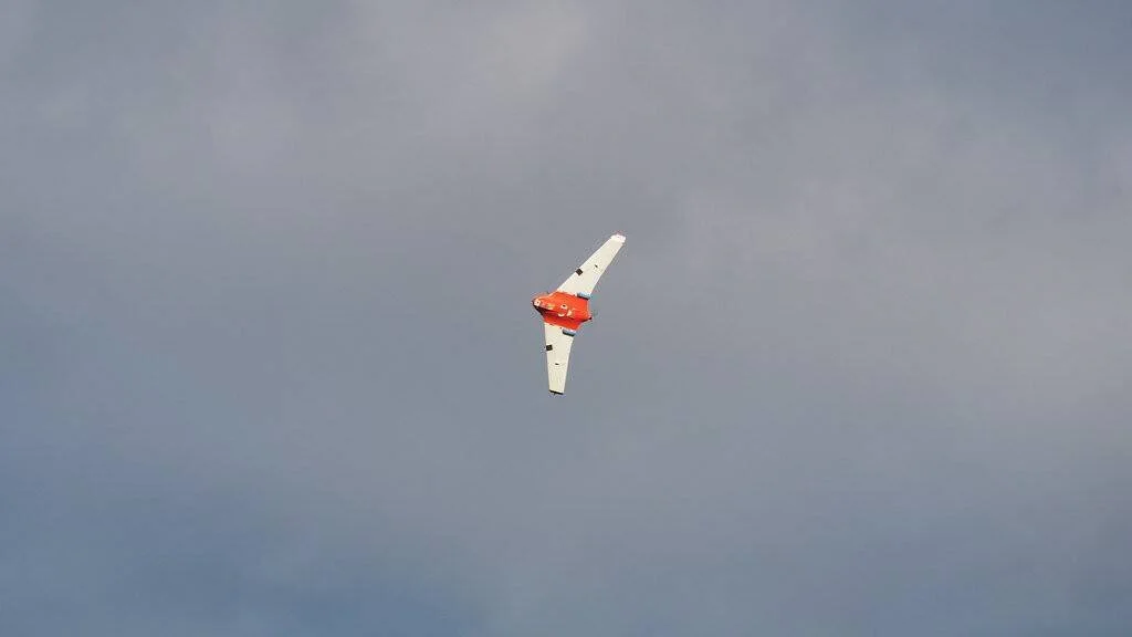 Dron de ala fija con cuerpo naranja y alas blancas volando en un cielo nublado.