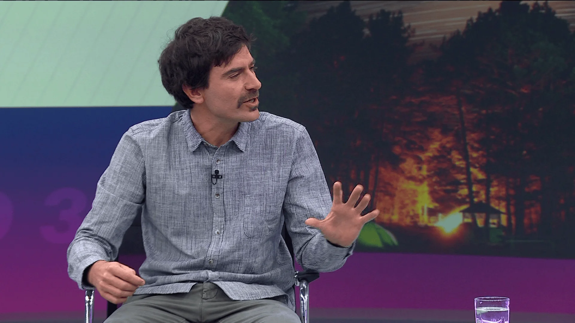 Hombre con bigote y camisa gris gesticula mientras habla en un estudio de televisión con imagen de incendio forestal al fondo.