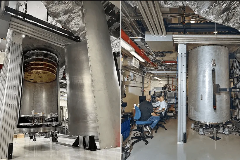 Dos técnicos trabajan frente a monitores junto a una gran cámara de vacío cilíndrica de metal en un laboratorio de pruebas.