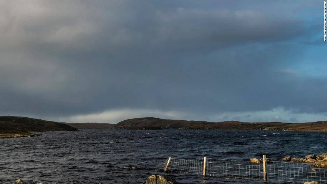 Lago de aguas oscuras agitadas bajo cielo tormentoso, con colinas bajas al fondo y una pequeña valla en la orilla.