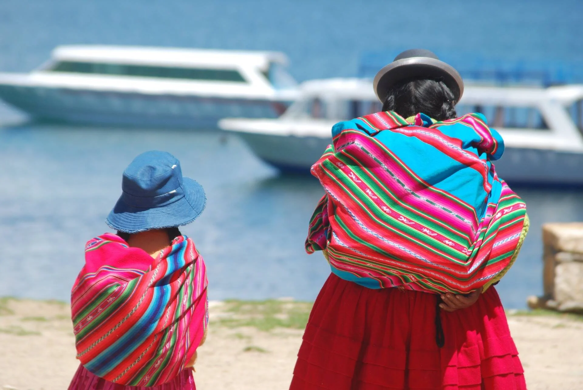 Dos mujeres con vestimenta tradicional andina de colores vivos observan embarcaciones en un lago, de espaldas a la cámara.
