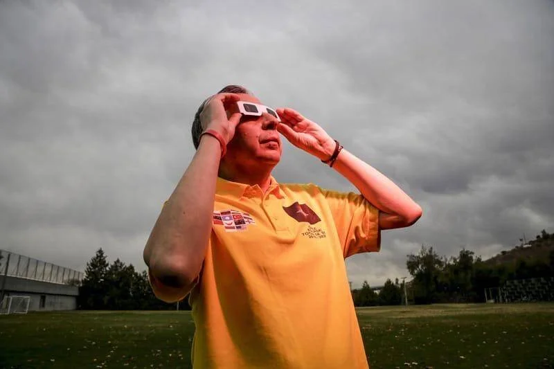 Hombre con remera amarilla usa anteojos de eclipse para mirar el cielo nublado en un campo abierto.