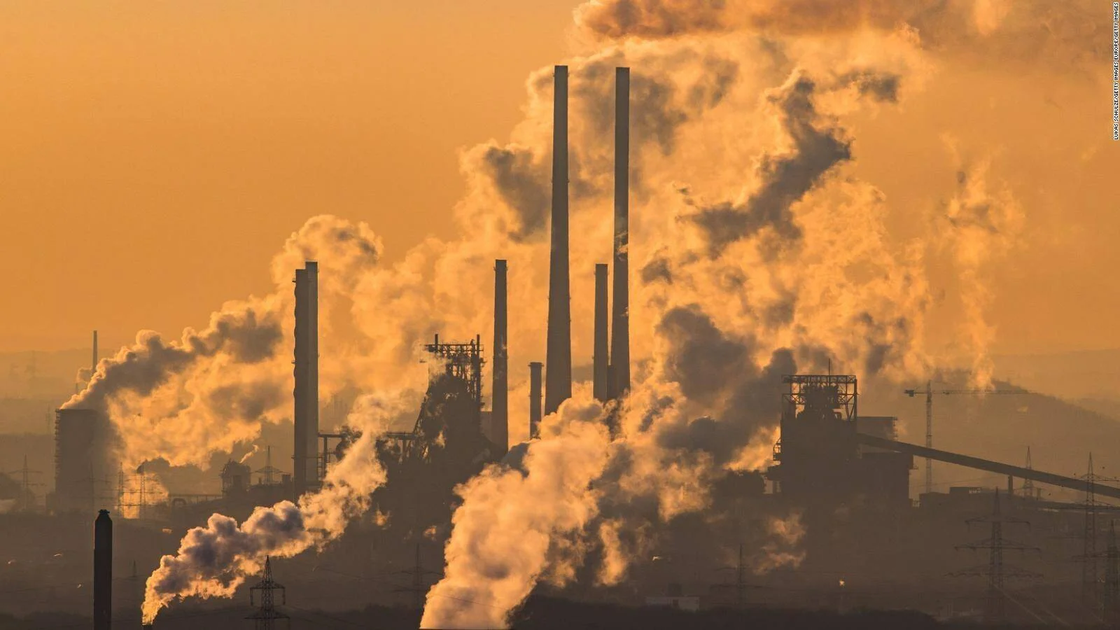 Complejo industrial con múltiples chimeneas expulsando grandes columnas de humo bajo un cielo naranja contaminado.