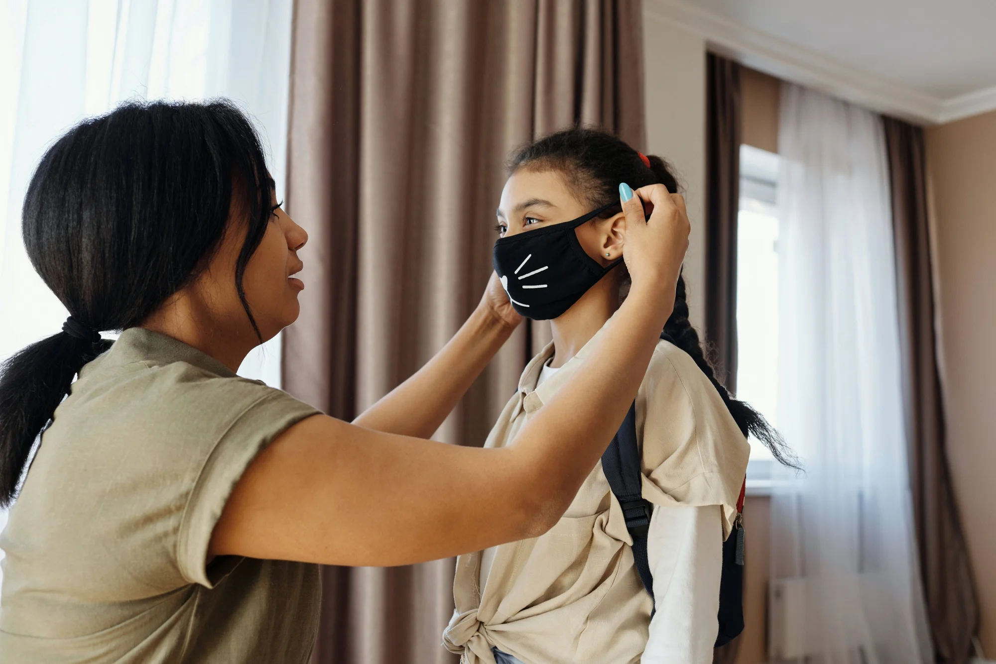 Una mujer ajusta una mascarilla negra decorada a una niña con mochila antes de salir de casa.