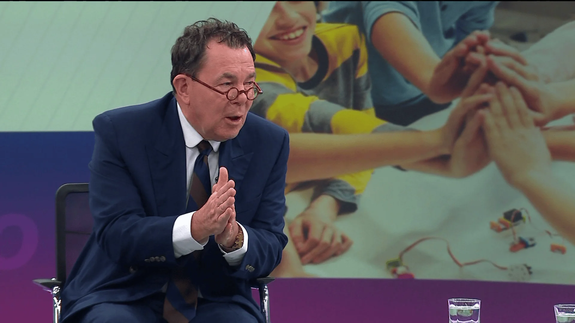 Hombre mayor con traje azul y gafas redondas habla gesticulando con las manos en un estudio de televisión.
