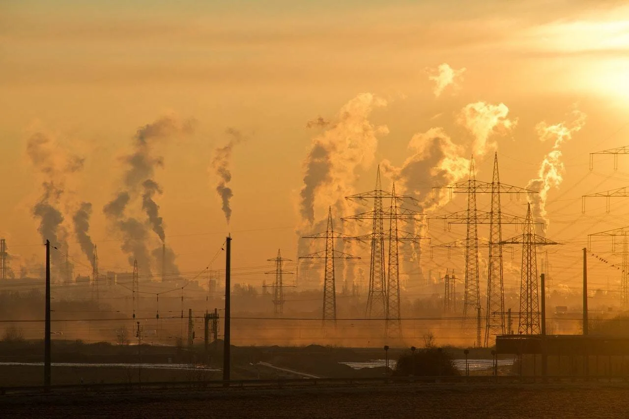 Zona industrial con múltiples chimeneas emitiendo humo y torres de alta tensión bajo un cielo anaranjado contaminado.