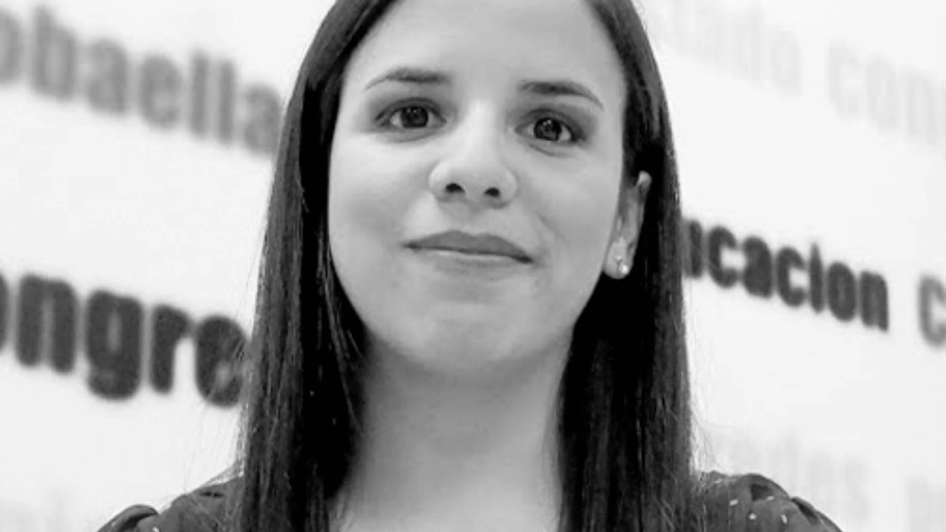 Mujer joven de cabello largo y liso posa frente a un fondo institucional con texto parcialmente visible.