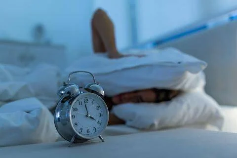 Persona en cama cubriéndose la cabeza con una almohada mientras un despertador analógico marca la hora en primer plano.
