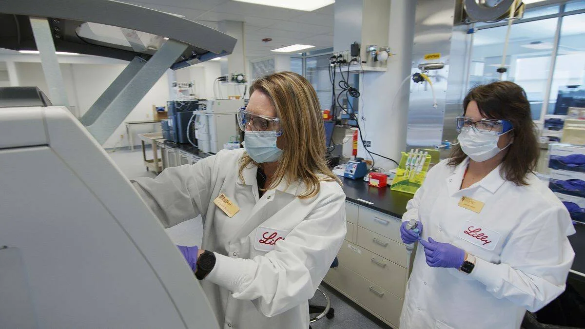 Dos científicas con batas blancas de Eli Lilly, mascarillas y guantes operan equipos en un laboratorio farmacéutico.