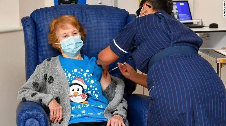 Una mujer mayor con mascarilla recibe una inyección de vacuna administrada por una enfermera en un centro de salud.