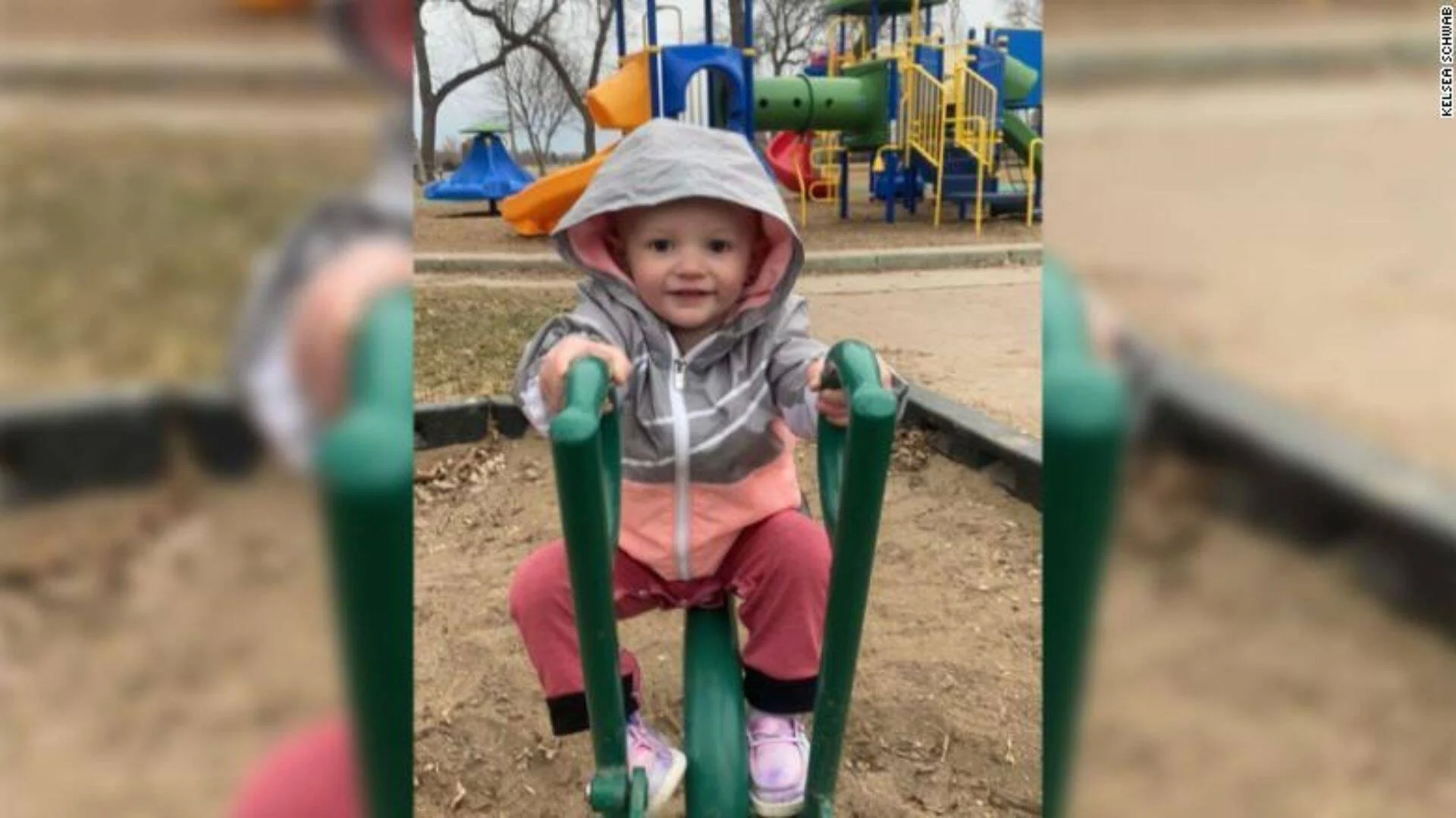 Bebé con sudadera gris y pantalón rosa montada en un juego de resorte en un parque infantil colorido.