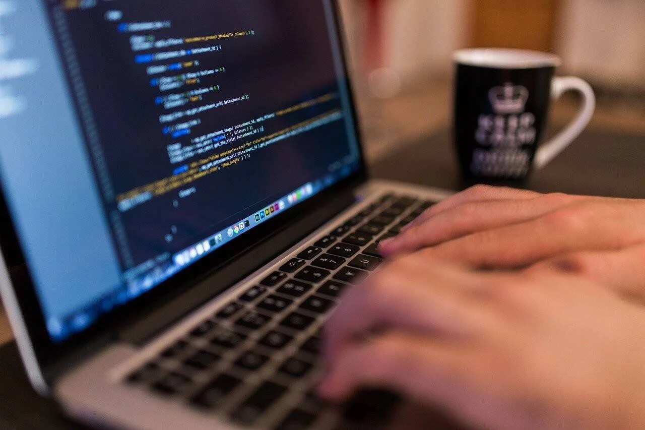 Manos escribiendo en el teclado de una laptop con código de programación en pantalla y una taza de café al fondo.