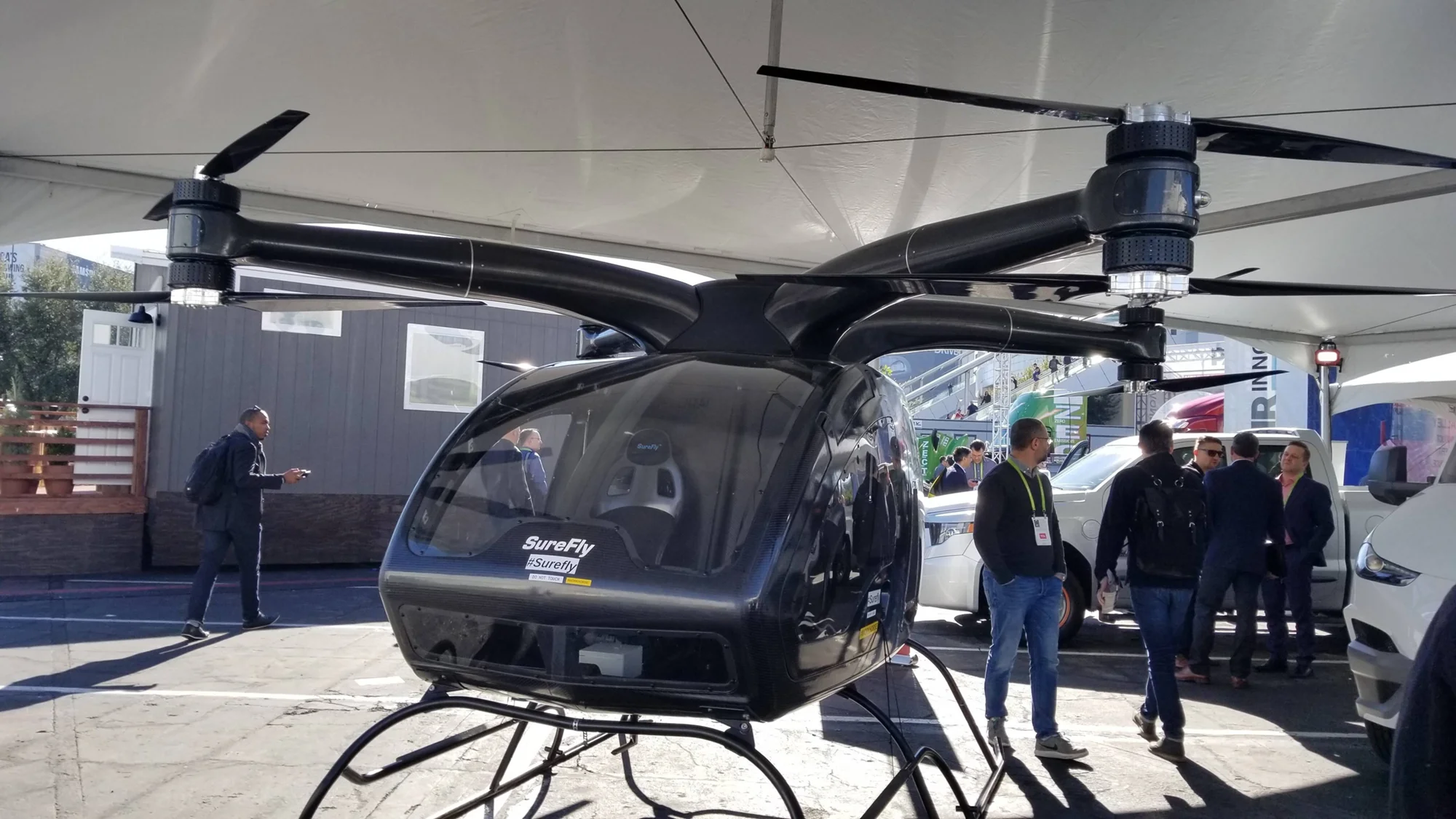 Helicóptero eléctrico multirrotor SureFly expuesto bajo una carpa rodeado de visitantes en una feria tecnológica.