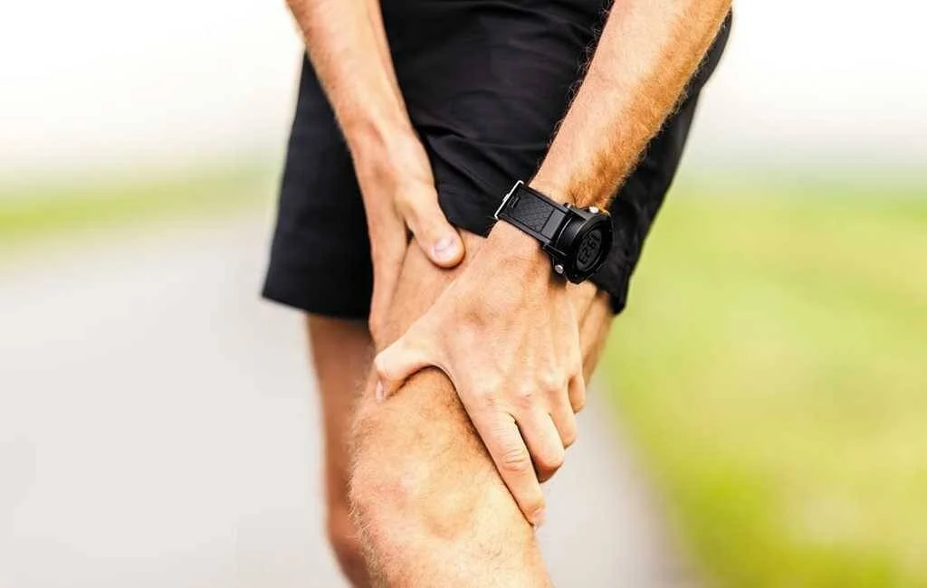 Corredor con ropa deportiva negra sujeta su rodilla con ambas manos, en señal de dolor o lesión durante el ejercicio.
