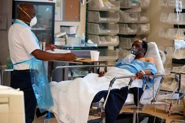 Profesional de salud con mascarilla y bata revisa documentos junto a paciente con máscara de oxígeno en cama hospitalaria.