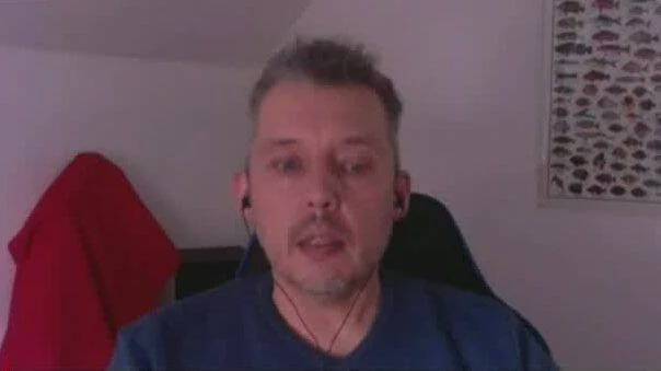 Hombre adulto con remera azul y auricular hablando frente a una cámara en una habitación.