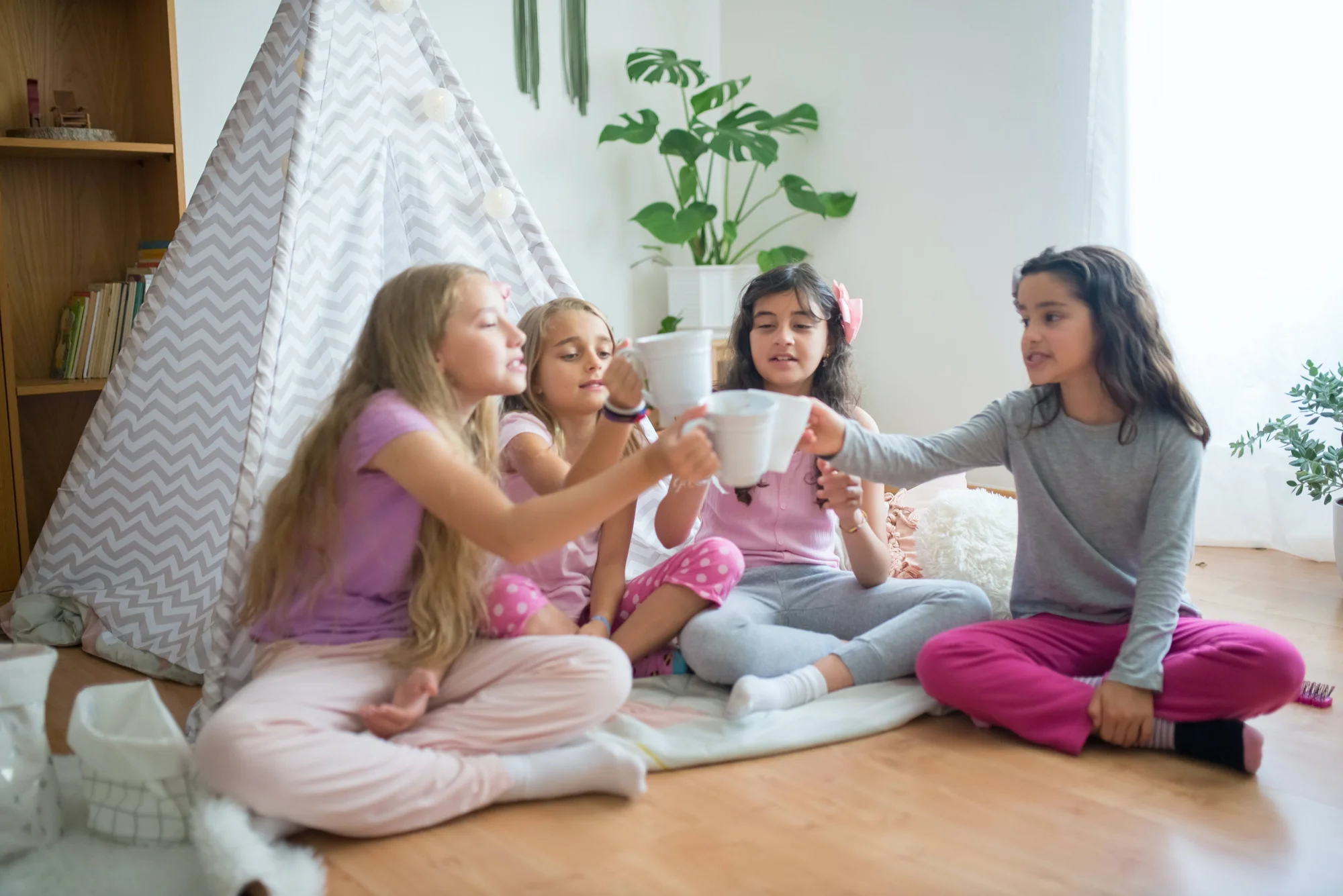 Cuatro niñas en pijama sentadas en el suelo brindan con tazas blancas frente a una carpa decorativa de juego.