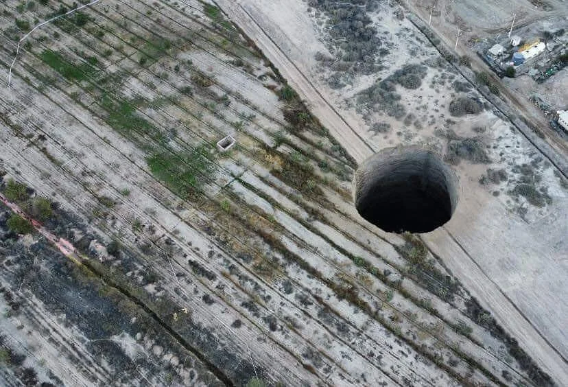 Vista aérea de un enorme socavón circular y profundo abierto en un terreno seco y árido, próximo a un camino de tierra.
