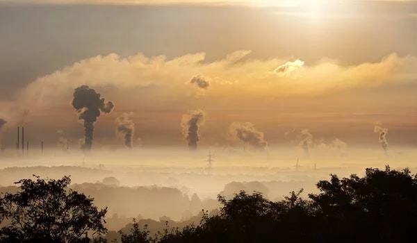 Zona industrial con múltiples chimeneas expulsando humo contaminante sobre un paisaje nublado al amanecer.