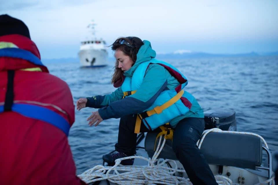 Una mujer con chaleco salvavidas azul trabaja a bordo de una pequeña embarcación con un buque de investigación al fondo.