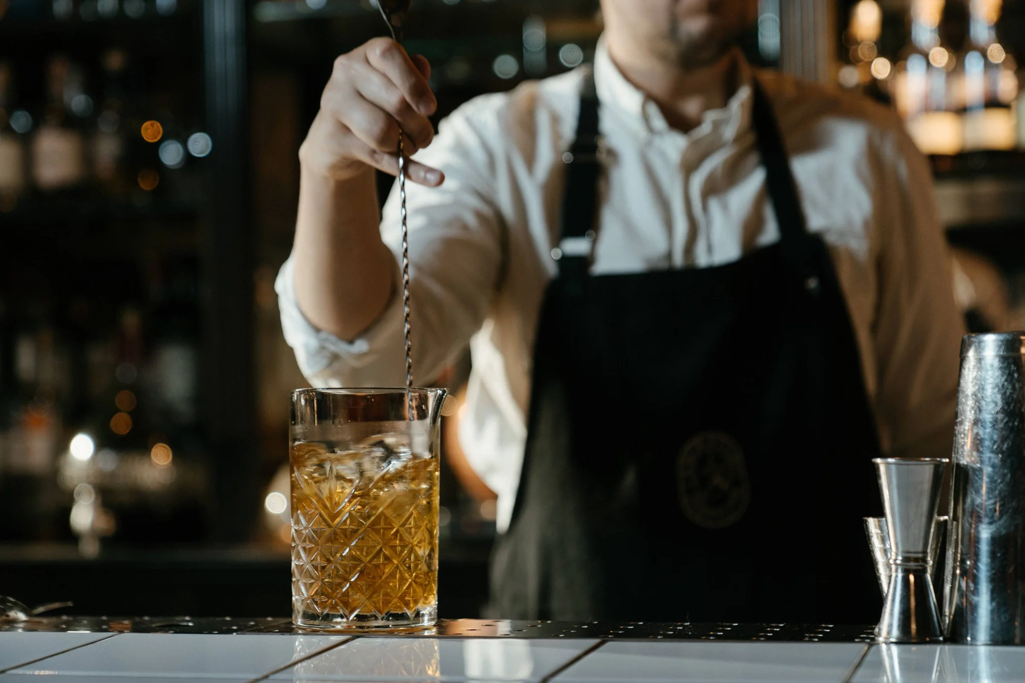 Bartender con delantal negro remueve un cóctel ambarino con hielo en vaso de cristal tallado sobre la barra.