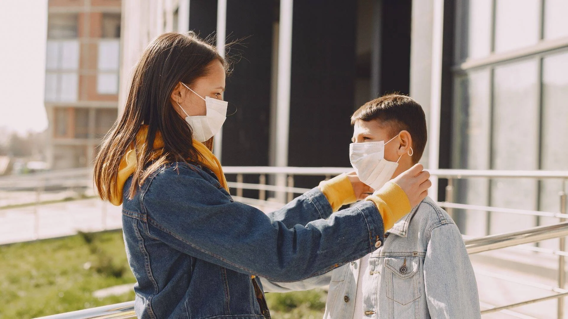 Una mujer con mascarilla ajusta el barbijo quirúrgico a un niño frente a un edificio al aire libre.
