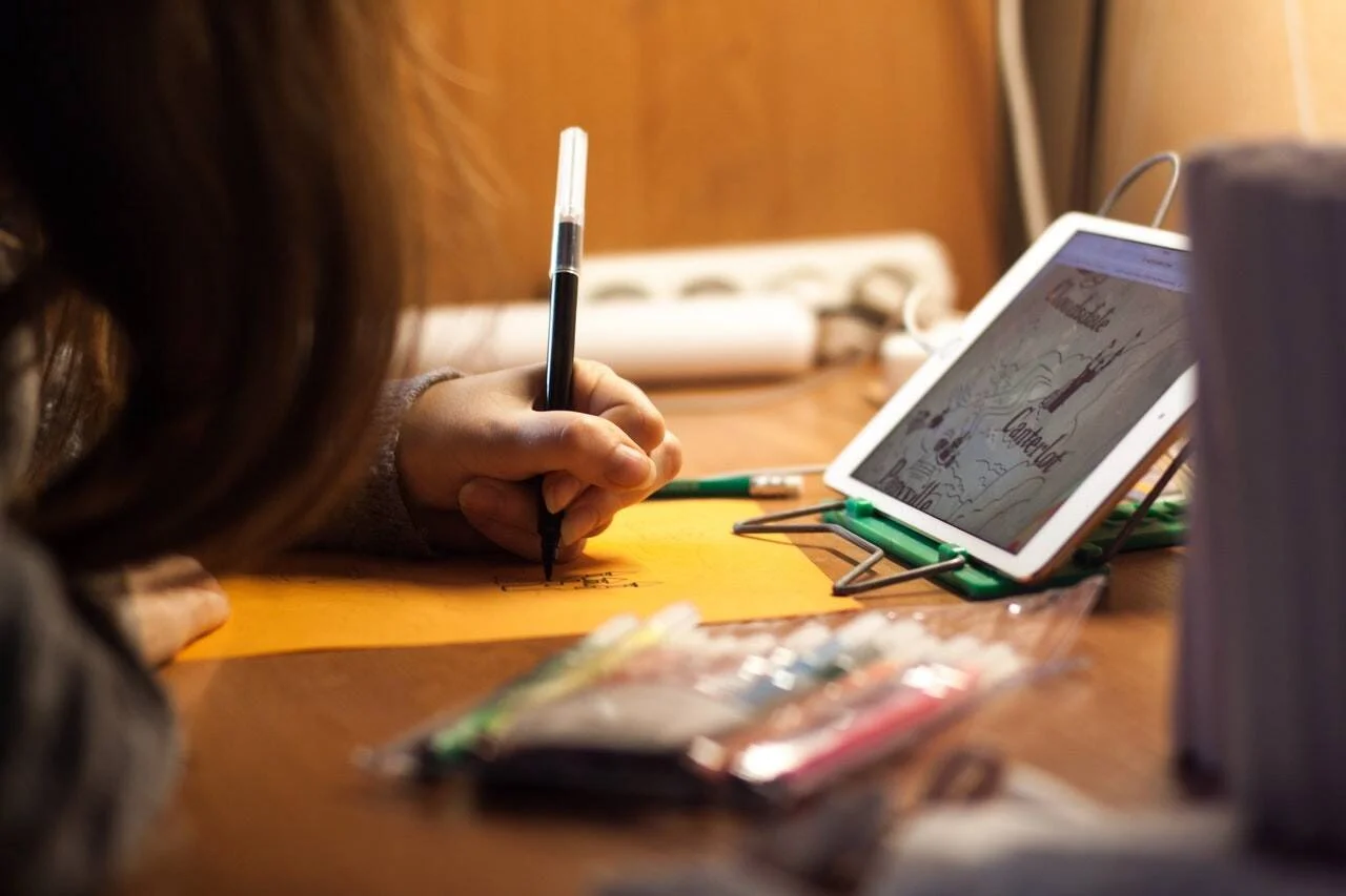 Una persona dibuja con pluma sobre papel naranja mientras usa una tablet como referencia visual en su escritorio.