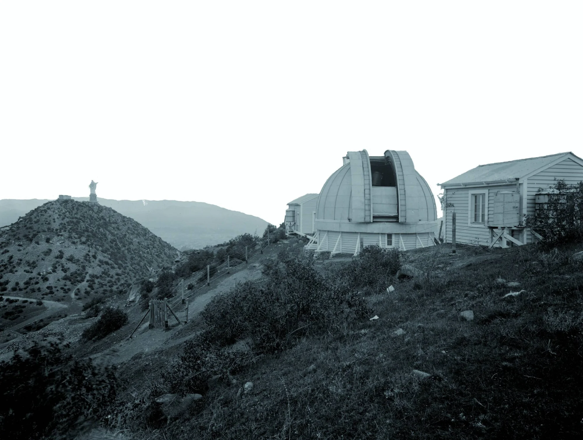 Edificio de observatorio astronómico con cúpula abierta en una colina, con una estatua religiosa visible en la cima de un cerro al fondo.